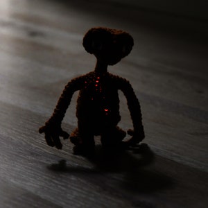 K&ouml;nnte beinhalten: Eine geh&auml;kelte braune Alienfigur mit leuchtend roten Akzenten. Das handgefertigte Spielzeug hat einen gro&szlig;en Kopf, lange Arme und einen kleinen K&ouml;rper. Die Figur steht auf einer Holzoberfl&auml;che, mit einem Schatten dahinter. Das Spielzeug ist eine Darstellung der Figur E.T.