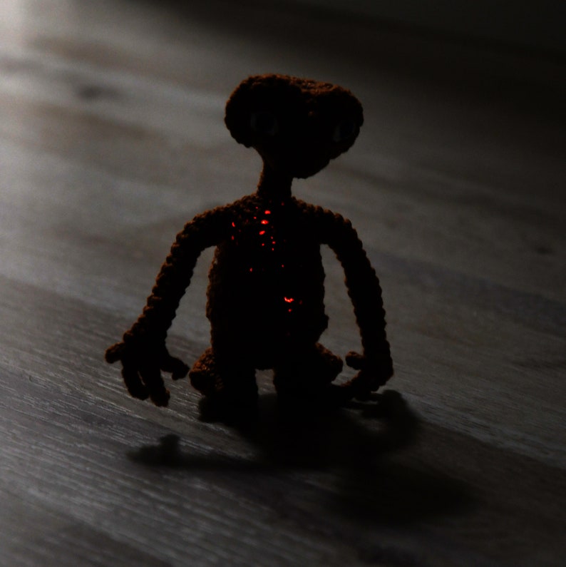 K&ouml;nnte beinhalten: Eine geh&auml;kelte braune Alienfigur mit leuchtend roten Akzenten. Das handgefertigte Spielzeug hat einen gro&szlig;en Kopf, lange Arme und einen kleinen K&ouml;rper. Die Figur steht auf einer Holzoberfl&auml;che, mit einem Schatten dahinter. Das Spielzeug ist eine Darstellung der Figur E.T.