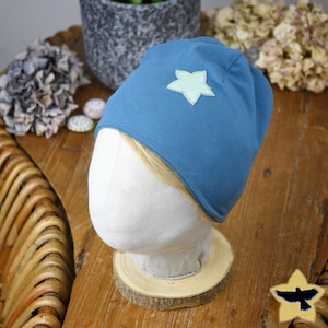 Könnte beinhalten: Ein blauer Beanie mit einem hellgrünen Stern-Applikation. Der Hut wird auf einem weißen Schaufensterkopf auf einem Holzsockel präsentiert. Der Hintergrund umfasst einen Holztisch, eine Topfpflanze und Trockenblumen.