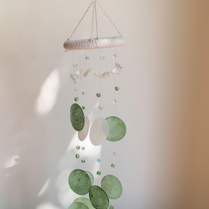 Può includere: Una giostrina sospesa con una parte superiore circolare avvolta in corda chiara, da cui scendono diversi fili da pesca trasparenti. I fili sono decorati con piccoli ciondoli a farfalla trasparenti, perline verdi e chiare e dischi circolari verdi e bianchi.