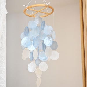 Puede incluir: Móvil colgante con un anillo de madera y una cascada de elementos iridiscentes en forma de disco en tonos azules y blancos. El móvil está suspendido de un cordón blanco. Un adorno delicado para guarderías o el hogar.