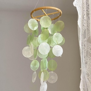 Op de afbeelding: Een hangende mobiel met een natuurlijke houten ring en strengen doorschijnende, ronde, groene en witte schelpschijven. De schijven variëren in grootte en tint, waardoor een gelaagd effect ontstaat. Woondecoratie.