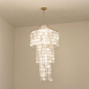 Puede incluir: Una lámpara de araña de varios niveles con múltiples filas de discos circulares de concha blanca translúcida suspendidos de un marco de metal dorado. La luminaria cuelga de un dosel de techo dorado a juego mediante una fina cadena.