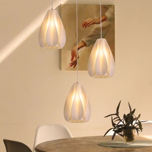 Puede incluir: Tres lámparas colgantes blancas con pantallas en forma de pétalos cuelgan del techo sobre una mesa de madera. Las luces están encendidas, proyectando un brillo cálido.