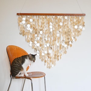 Puede incluir: Un colgante de pared decorativo hecho de numerosas piezas pequeñas y redondas de concha iridiscente. Las conchas están suspendidas de una barra de madera, creando una forma triangular en cascada. Un gato está sentado en una silla naranja en primer plano.
