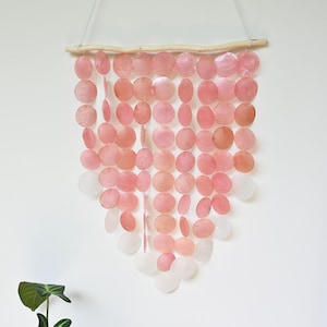 Op de afbeelding: Een wandhanger met meerdere verticale strengen van ronde, doorschijnende roze en witte capiz-schelpen die aan een horizontale houten tak hangen. De schelpen zijn in een V-vorm gerangschikt met een wit touwtje aan de tak bevestigd.
