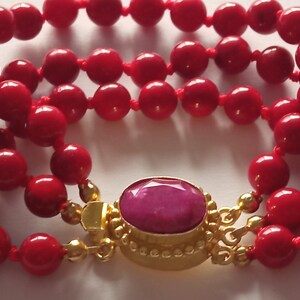 Pode incluir: Um bracelete de contas de coral vermelho com um fecho dourado e uma gema rosa no centro.