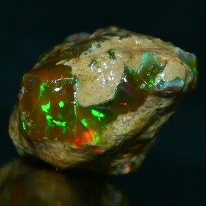 Puede incluir: Una piedra preciosa de ópalo en bruto con un brillo iridiscente verde y rojo. El ópalo está incrustado en una matriz marrón.