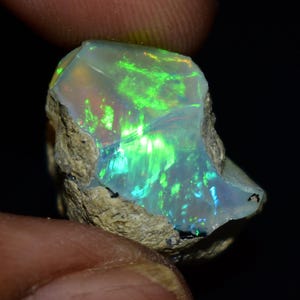 Könnte beinhalten: Ein roher Opal-Edelstein mit einem lebendigen Farbspiel. Der Stein zeigt Blitze von Grün, Blau und Orange in einer durchscheinenden, milchig-weißen Basis. Der Opal ist vor einem dunklen Hintergrund platziert, was seine irisierenden Eigenschaften hervorhebt.