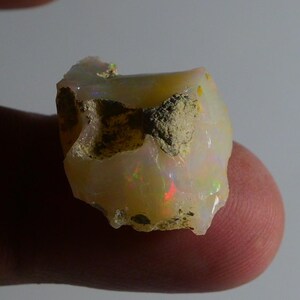 Könnte beinhalten: Ein roher Opal-Edelstein, überwiegend cremeweiß, mit roten, grünen und gelben irisierenden Blitzen. Der Stein hat eine raue, strukturierte Oberfläche mit Bereichen aus beiger Matrix. Ein natürlicher, ungeschliffener Edelstein.