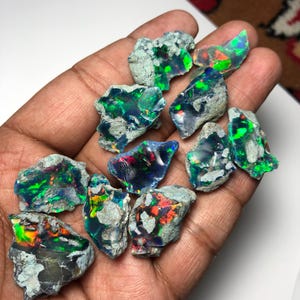 Könnte beinhalten: Eine Sammlung roher Opal-Edelsteine, die lebhafte Farbblitze wie Grün, Blau, Rot und Gelb zeigen. Die Opale sind vor einem hellen Hintergrund platziert, der ihre natürlichen Texturen und irisierenden Eigenschaften hervorhebt. Die Opale sind in verschiedenen Größen und Formen.