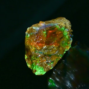 May include: A rough, natural opal gemstone with a green, orange, and red iridescent sheen. The opal is set in a matrix of brown rock.