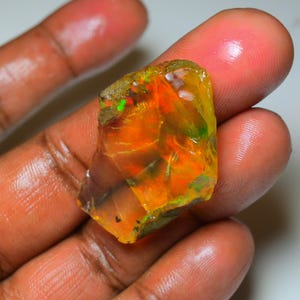 Puede incluir: Una piedra preciosa de ópalo en bruto con vibrantes tonos naranja, amarillo y verde. La piedra tiene una cualidad translúcida, con patrones internos visibles y un exterior rugoso y natural. El ópalo se sostiene entre los dedos, mostrando su tamaño y variaciones de color.