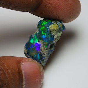 Könnte beinhalten: Ein roher Opal-Edelstein mit lebendigen Farben von Grün, Blau und Violett. Der Stein hat eine dunkle Basis mit schillernden Farbtönen. Der Opal wird zwischen zwei Fingern gehalten, was seine natürliche, unpolierte Textur zeigt.