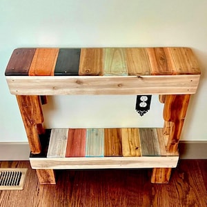 May include: A rustic wooden console table with a multi-colored top made of reclaimed wood planks. The table has a lower shelf and sturdy legs, showcasing a natural wood grain finish. The table is 48 inches long.