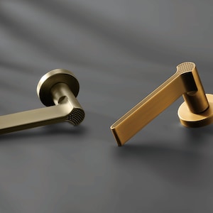 May include: Two gold-colored door handles. The handles have a modern design with a rectangular lever and a circular base. The end of the lever has a textured pattern. The handles are made of metal and are displayed on a dark gray surface.