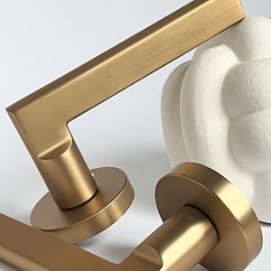 May include: Two gold door handles with a square design. The handles are made of metal and have a brushed finish.