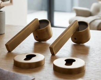 May include: Two brass door handles with matching circular escutcheons featuring keyhole cutouts. The hardware has a brushed metallic finish and is displayed alongside a metal caliper tool on a wooden surface.