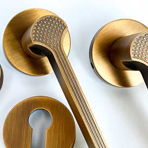 Peut inclure: Un ensemble de trois poignées de porte dorées avec un motif géométrique. Les poignées ont une base ronde et un levier long et incurvé.