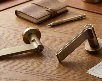 May include: Two brass door handles with a matte finish and a textured, knurled pattern on the top edge. The handles feature a cylindrical base and a flat, rectangular lever design. The metal surface has a muted, brushed gold appearance.