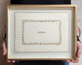 May include: A white wooden serving tray with a gold trim and a gold leaf design. The tray has two handles on the sides.