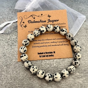 Puede incluir: Un brazalete hecho de cuentas de jaspe dalmata blancas y negras. El brazalete está sobre una tarjeta marrón con el texto "Dalmatian Jasper" e información sobre las propiedades de la piedra. La tarjeta también dice "Handmade in Warwickshire, UK by Crystal Craft Jewellery."