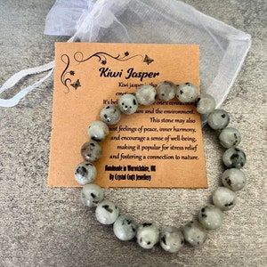 May include: A green and black kiwi jasper beaded bracelet with a brown card that reads "Kiwi Jasper" and includes information about the stone's properties. The bracelet is handmade in Warwickshire, UK by Crystal Craft Jewellery.