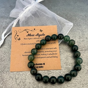 May include: A green moss agate bracelet with 14 beads. The bracelet is on a brown card with the text "Moss Agate" and information about the stone's properties.
