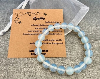 Könnte beinhalten: Ein hellblaues Opalit-Armband mit weißen Streifen auf jeder Perle. Das Armband liegt auf einer braunen Karte mit dem Text "Opalite" und Informationen über die Eigenschaften des Steins.