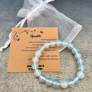 Puede incluir: Un brazalete de ópalo azul claro con rayas blancas en cada cuenta. El brazalete está sobre una tarjeta marrón con el texto "Opalite" e información sobre las propiedades de la piedra.