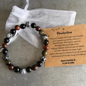 May include: A bracelet made of mixed-colour gemstone beads, including black, red, grey, and green. The bracelet is presented with a small white bag and a brown tag with the word "Protection" and text describing the stones' properties.