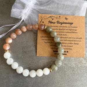 Könnte beinhalten: Ein Perlenarmband mit weißen, pfirsichfarbenen und grünen Steinen, akzentuiert mit goldfarbenen Perlen. Das Armband wird mit einem kleinen weißen Kordelzugbeutel und einer Karte mit der Aufschrift "New Beginnings" präsentiert.