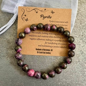 K&ouml;nnte beinhalten: Ein Armband mit dunklen, metallisch aussehenden Pyrit- und rosa Edelsteinen. Das Armband wird mit einer Karte mit der Aufschrift "Pyrit" und einem wei&szlig;en Organza-Geschenkbeutel pr&auml;sentiert.