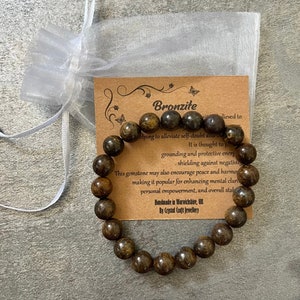 Puede incluir: Un brazalete de piedra de bronzita marrón con una tarjeta que dice "Bronzite" y describe las propiedades de la piedra. La tarjeta dice "ayudar a aliviar la duda propia y la ansiedad. Se cree que proporciona energía de conexión a tierra y protección, protegiendo contra la negatividad. Esta piedra preciosa también puede fomentar la paz y la armonía, lo que la hace popular para mejorar la claridad mental, el empoderamiento personal y la estabilidad general. Hecho a mano en Warwickshire, Reino Unido por Crystal Craft Jewellery."