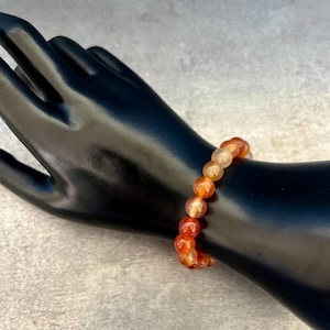 May include: A beaded bracelet featuring orange and amber-colored gemstones. The bracelet is made of small, round beads strung together. The gemstones have a translucent quality, and the bracelet is displayed on a black mannequin hand.