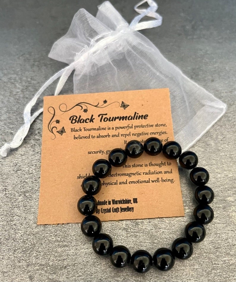Op de afbeelding: Zwarte toermalijn armband gemaakt met gepolijste zwarte kralen. De armband ligt op een bruine kaart met de tekst: "Zwarte toermalijn is een krachtige beschermende steen, waarvan wordt aangenomen dat hij negatieve energie&euml;n absorbeert en afstoot. Deze steen wordt verondersteld elektromagnetische straling en fysiek en emotioneel welzijn te beschermen. Gemaakt in Warwickshire, VK door Crystal Craft Jewellery."