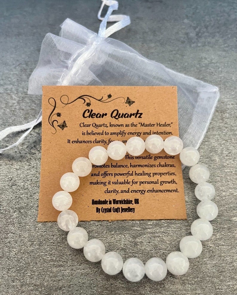 May include: A clear quartz bracelet with large, round beads. The bracelet is displayed with a small, sheer white drawstring bag and a brown card with the text "Clear Quartz" and product information. The bracelet is handmade in Warwickshire, UK.