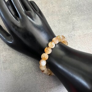 May include: A bracelet made of golden-yellow and translucent citrine gemstones. The bracelet is shown on a black hand-shaped display against a grey background. The beads are approximately 1 cm in diameter.