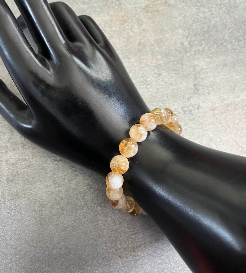 May include: A bracelet made of golden-yellow and translucent citrine gemstones. The bracelet is shown on a black hand-shaped display against a grey background. The beads are approximately 1 cm in diameter.