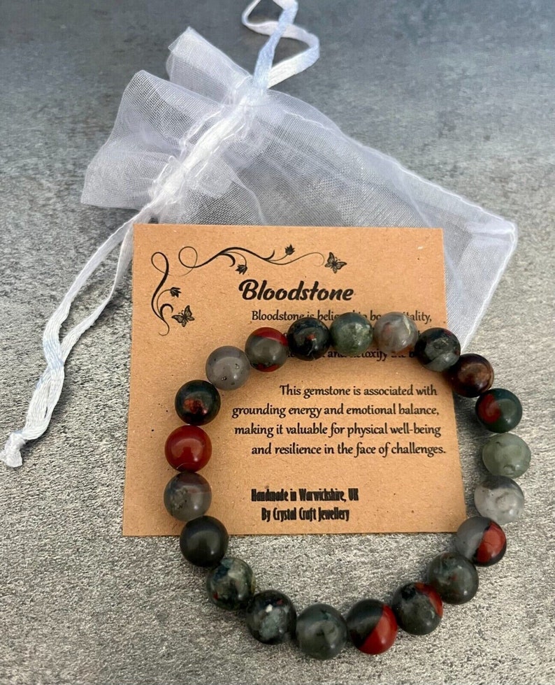 K&ouml;nnte beinhalten: Ein Blutstein-Armband mit roten und gr&uuml;nen Steinen. Das Armband liegt auf einer braunen Karte mit dem Text "Bloodstone" und "Dieser Edelstein ist mit erdendem Energie und emotionalem Gleichgewicht verbunden, was ihn wertvoll f&uuml;r das k&ouml;rperliche Wohlbefinden und die Widerstandsf&auml;higkeit im Angesicht von Herausforderungen macht." Die Karte tr&auml;gt au&szlig;erdem den Text "Handgefertigt in Warwickshire, UK von Crystal Craft Jewellery".
