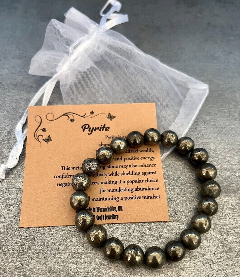 May include: A brown card with the text "Pyrite" and a description of the stone's properties. A bracelet made of round, dark gray pyrite beads is on top of the card.