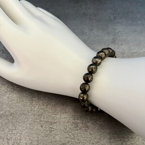 May include: A bracelet made of round, dark gray pyrite beads. The bracelet is on a white mannequin wrist.