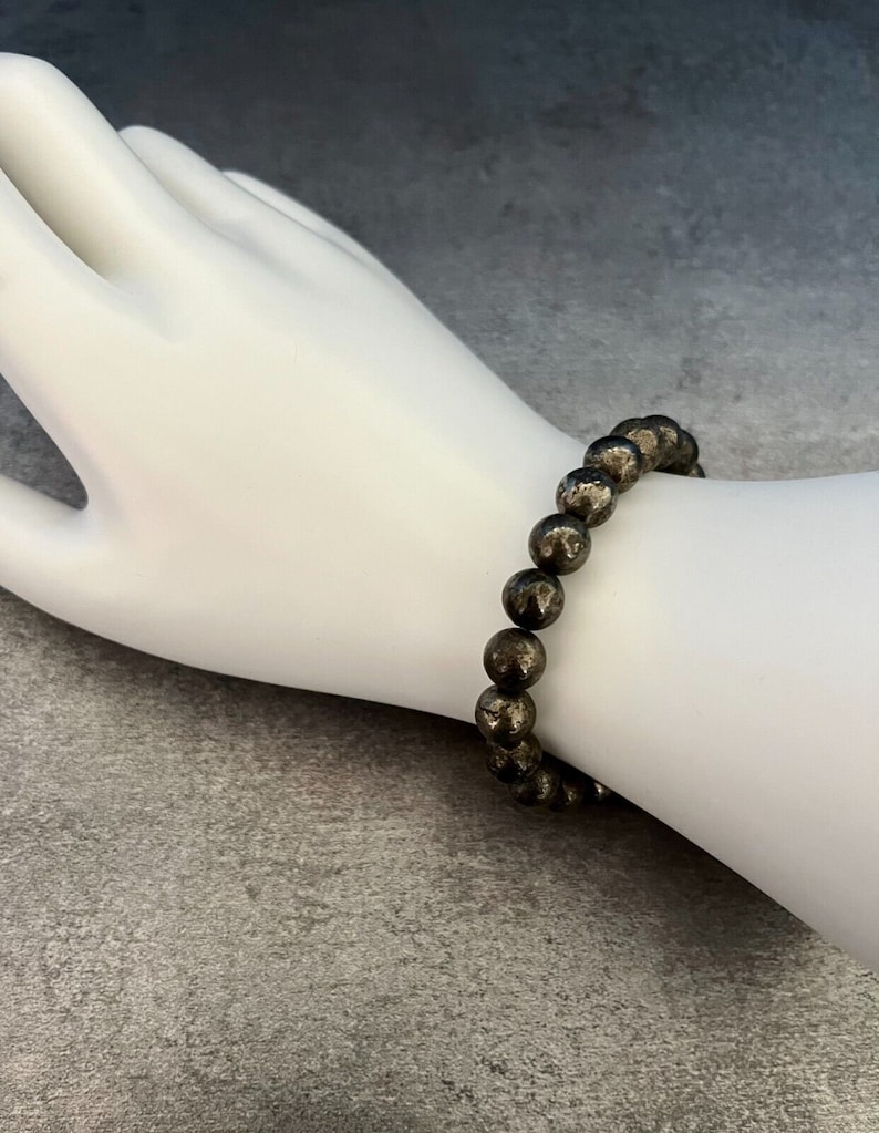 May include: A bracelet made of round, dark gray pyrite beads. The bracelet is on a white mannequin wrist.