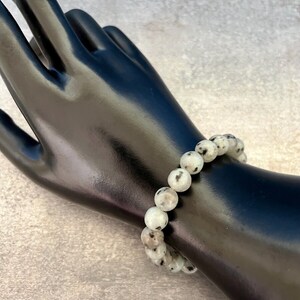 May include: A white and black speckled stone beaded bracelet. The bracelet is made of round beads and is worn on a mannequin's wrist.