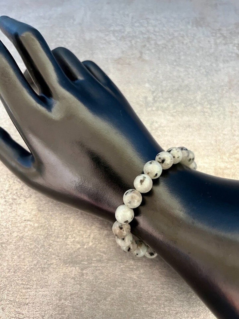 May include: A white and black speckled stone beaded bracelet. The bracelet is made of round beads and is worn on a mannequin's wrist.