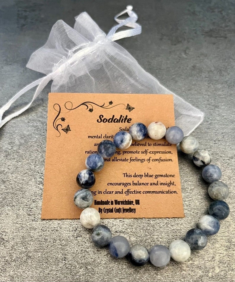 May include: A blue and white sodalite gemstone bracelet with a brown card that reads "Sodalite" and describes the stone's properties. The card also says "This deep blue gemstone encourages balance and insight, bringing in clear and effective communication."