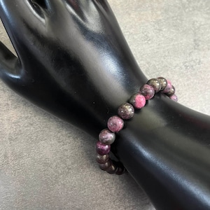 K&ouml;nnte beinhalten: Ein Armband mit abwechselnd dunkelgrauen und rosaroten runden Perlen. Das Armband wird auf einem schwarzen, handf&ouml;rmigen Schmuckst&auml;nder vor grauem Hintergrund pr&auml;sentiert. Die Perlen haben einen Durchmesser von etwa 1,3 cm.