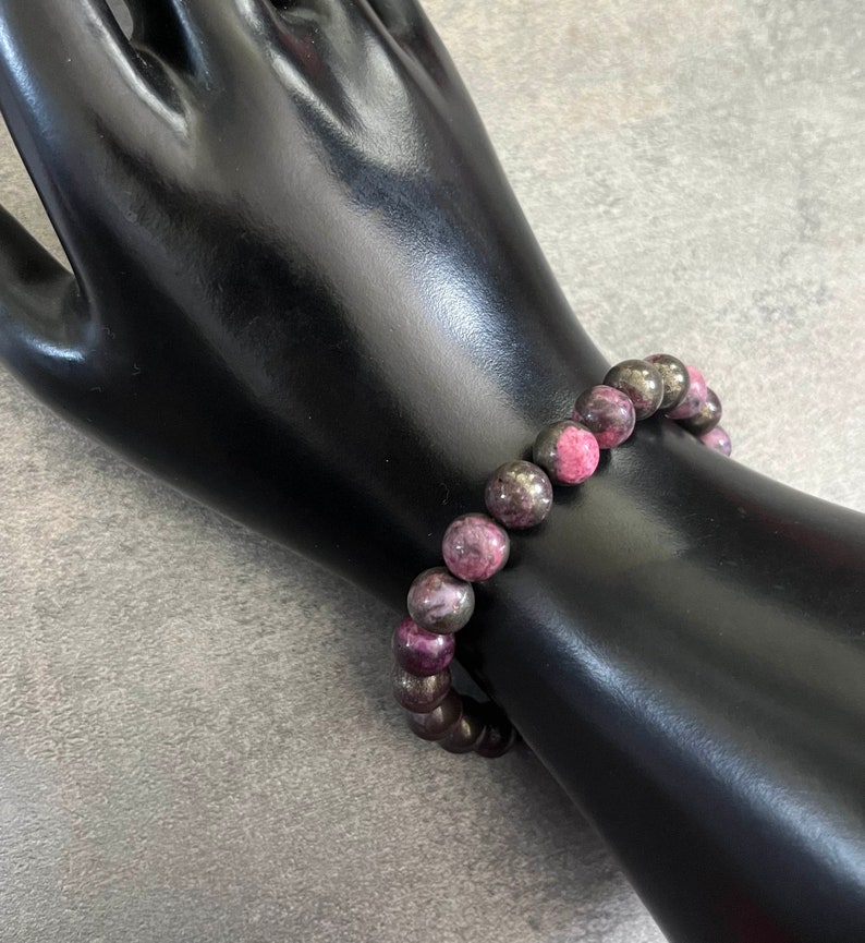 K&ouml;nnte beinhalten: Ein Armband mit abwechselnd dunkelgrauen und rosaroten runden Perlen. Das Armband wird auf einem schwarzen, handf&ouml;rmigen Schmuckst&auml;nder vor grauem Hintergrund pr&auml;sentiert. Die Perlen haben einen Durchmesser von etwa 1,3 cm.