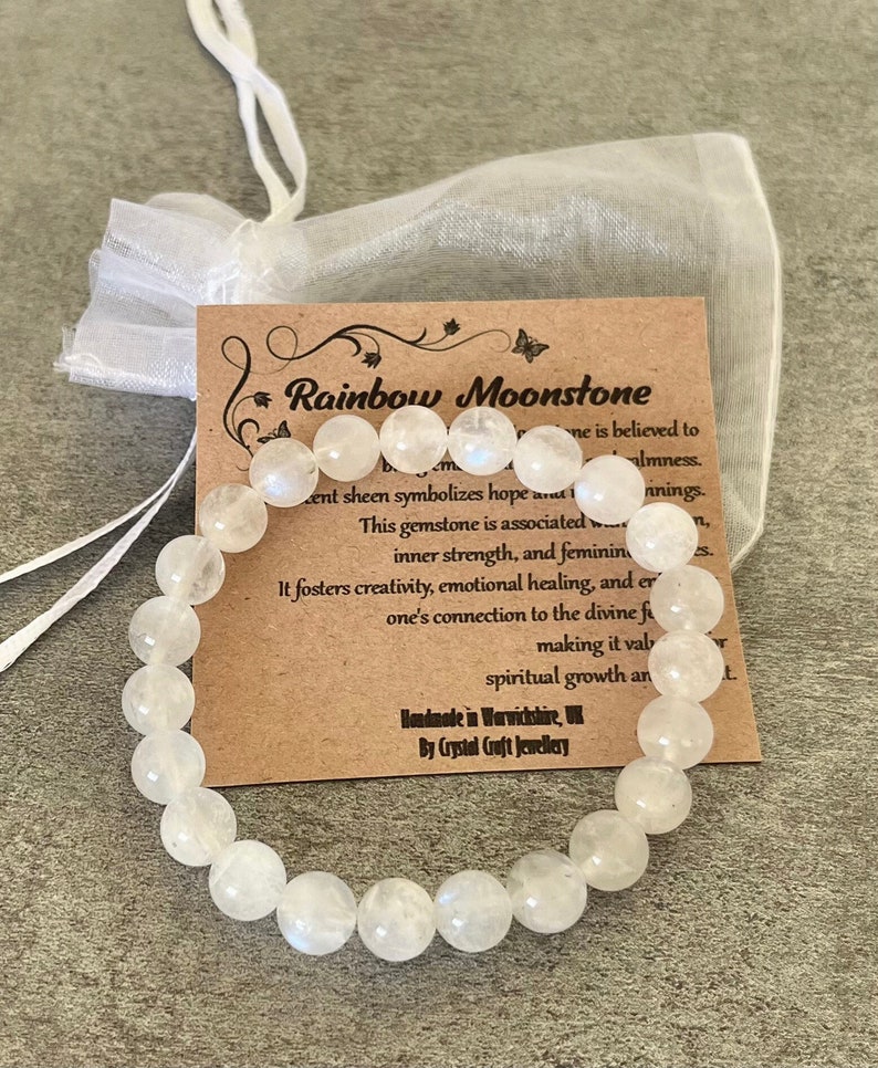 May include: A white rainbow moonstone bracelet with a brown card that reads "Rainbow Moonstone" and includes information about the gemstone's properties. Handmade in Warwickshire, UK by Crystal Craft Jewellery.