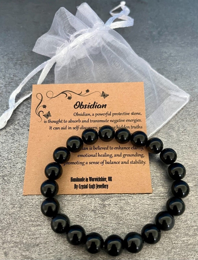 May include: Black obsidian gemstone bracelet with a brown card that reads "Obsidian, a powerful protective stone, is thought to absorb and transmute negative energies. It can aid in self-discovery, revealing hidden truths. Obsidian is believed to enhance clarity, emotional healing, and grounding, promoting a sense of balance and stability. Handmade in Warwickshire, UK By Crystal Craft Jewellery"